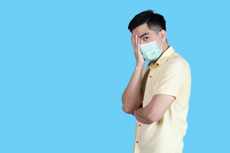 Stressful COVID-19 Coronavirus portrait handsome young asian 
man wearing yellow shirt and mask protection from covid 19 isolated on blue background in studio. Asian man people. COVID-19 concept.の写真素材