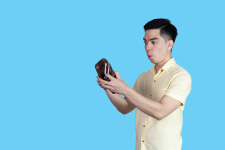 Portrait Handsome young asian man wearing a yellow shirt surpise money in the wallet isolated on blue background. Asia people.の写真素材