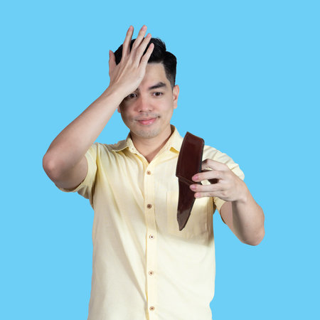 Portrait of a handsome young Asian man wearing a yellow shirt stressed because of an empty wallet, isolated on a blue background.の写真素材