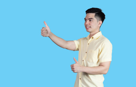 Portrait handsome young asian man wearing a yellow shirt showing thumbs up sign isolated on blue background. Asian man people. business success concept.の写真素材
