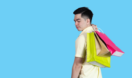 Portrait handsome young asian man wearing yellow cheerful glad content guy carrying new things colorful packages on blue background in studio. Asian man people.の写真素材