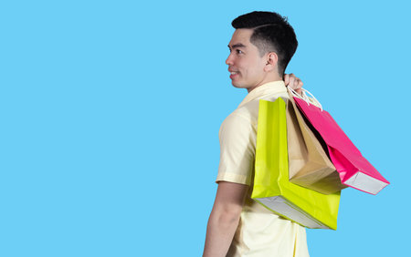 Portrait handsome young asian man wearing yellow cheerful glad content guy carrying new things colorful packages on blue background in studio. Asian man people.の写真素材