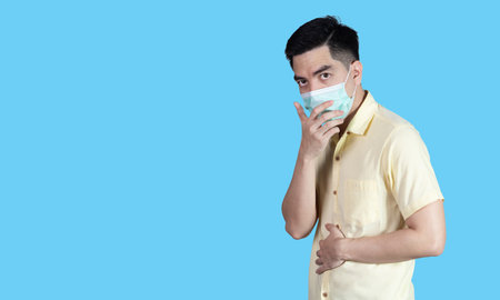 COVID-19 portrait handsome young asian man wearing yellow shirt and sneezing mask protection isolated on blue background in studio. Asian man people. COVID-19 concept.の写真素材