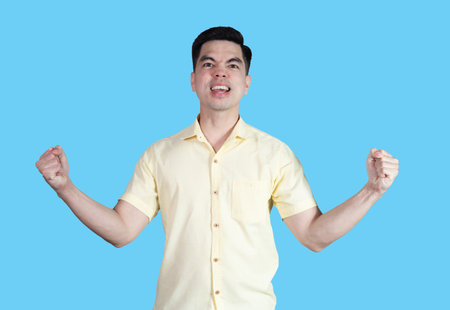 Portrait handsome young asian man wearing a yellow shirt showing excitement or celebrating  sign isolated on blue background. Asian man people. business success concept.の写真素材