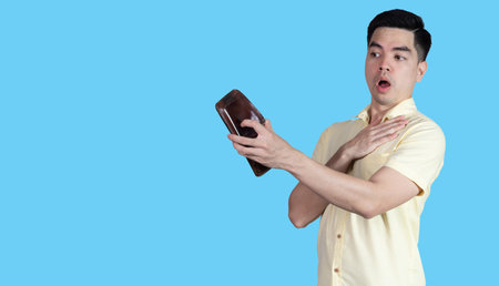 Portrait of a young Asian man wearing a yellow shirt, stressed because of an empty wallet, isolated on a blue background.の写真素材