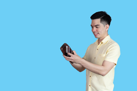 Portrait Handsome young asian man wearing a yellow shirt feeling happy with money in the wallet isolated on blue background.の写真素材