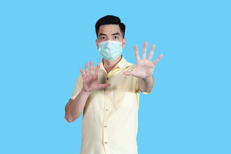 Portrait handsome young asian man wearing yellow shirt and mask protection isolated on blue background in studio. Asian man people.の写真素材