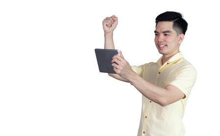 Portrait of a handsome young Asian man wearing a yellow shirt, holding a smart phone or tablet, and celebrating or feeling successful, isolated on a blue background.の写真素材