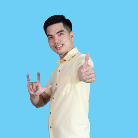 Portrait handsome young asian man wearing a yellow shirt showing thumbs up sign isolated on blue background. Asian man people. business success concept.の写真素材