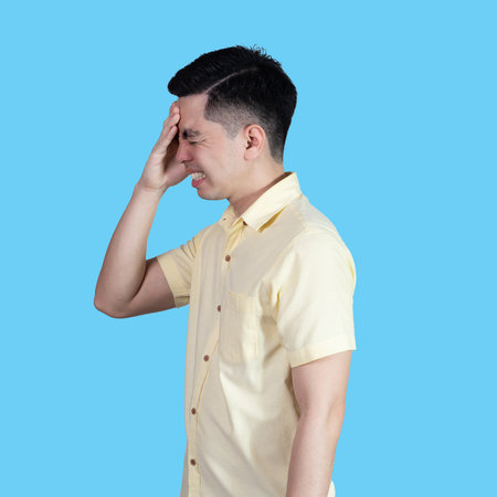 Portrait of a handsome young Asian man wearing a yellow shirt, stressed and showing signs of anxiety, isolated on a blue background in a studio.の写真素材