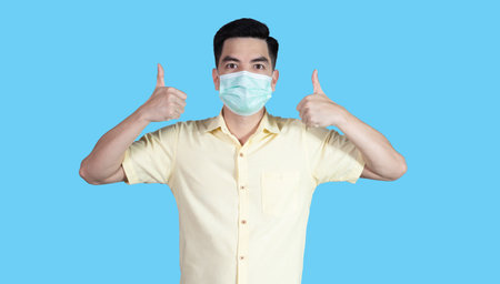 COVID-19 Coronavirus portrait handsome young asian man wearing yellow shirt and mask protection from covid 19 and thumbs up sign isolated on blue background in studio. Asian people. COVID-19 concept.の写真素材