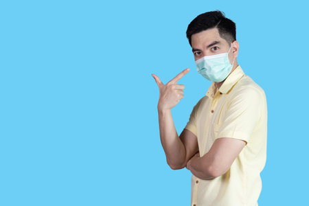 COVID-19 Coronavirus portrait handsome young asian man wearing yellow shirt and mask protection from covid 19 and thumbs up sign isolated on blue background in studio. Asian people. COVID-19 concept.の写真素材