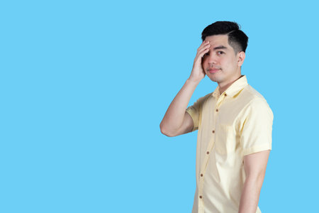 Portrait of a young Asian man wearing a yellow shirt, stressed and anxious, isolated on a blue background in a studio. Asian man.の写真素材
