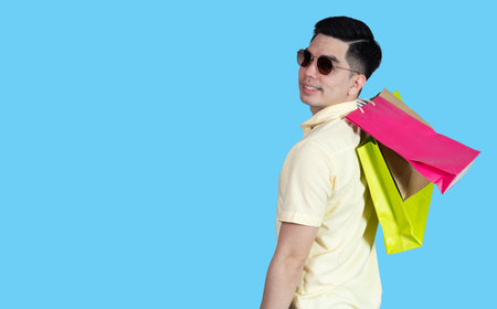 Portrait handsome young asian man wearing yellow cheerful glad content guy carrying new things colorful packages on blue background in studio. Asian man people.の写真素材