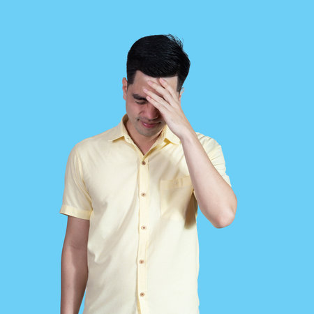 Portrait of a young Asian man wearing a yellow shirt, looking stressed and anxious, isolated on a blue background in a studio.の写真素材
