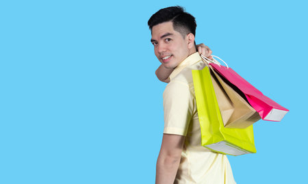 Portrait handsome young asian man wearing yellow cheerful glad content guy carrying new things colorful packages on blue background in studio. Asian man people.の写真素材