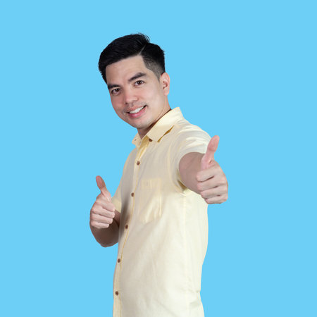 Portrait handsome young asian man wearing a yellow shirt showing thumbs up sign isolated on blue background. Asian man people. business success concept.の写真素材