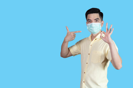 COVID-19 Coronavirus portrait handsome young asian man wearing yellow shirt and mask protection from covid 19 and thumbs up sign isolated on blue background in studio. Asian people. COVID-19 concept.の写真素材