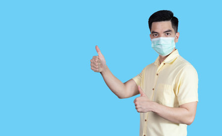 COVID-19 Coronavirus portrait handsome young asian man wearing yellow shirt and mask protection from covid 19 and thumbs up sign isolated on blue background in studio. Asian people. COVID-19 concept.の写真素材