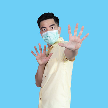 Portrait of a handsome young Asian man wearing a yellow shirt and a mask for protection, isolated on a blue background in a studio. Asian man. Concept of protection.の写真素材