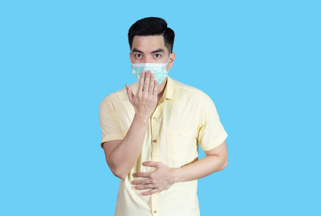 COVID-19 portrait handsome young asian man wearing yellow shirt and sneezing mask protection isolated on blue background in studio. Asian man people. COVID-19 concept..の写真素材