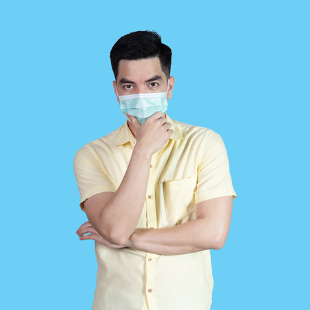 COVID-19 Coronavirus portrait handsome young asian man wearing yellow shirt and mask protection from covid 19 isolated on blue background in studio. Asian man people. COVID-19 concept.の写真素材