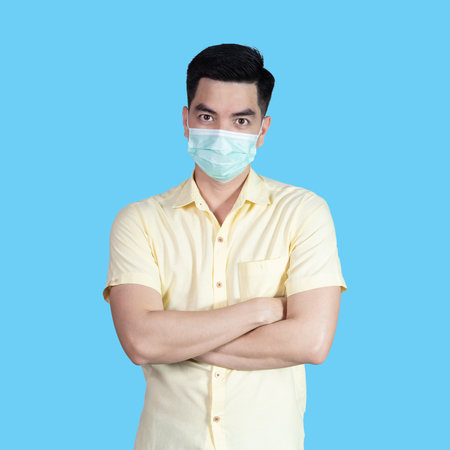 COVID-19 Coronavirus portrait handsome young asian man wearing yellow shirt and mask protection from covid 19 isolated on blue background in studio. Asian man people. COVID-19 concept.の写真素材
