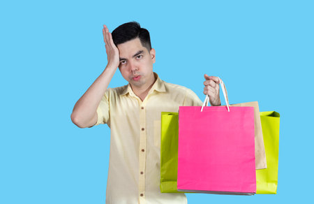 Portrait of a handsome young Asian man wearing a yellow shirt, carrying colorful packages on a blue background in a studio. Asian man.の写真素材