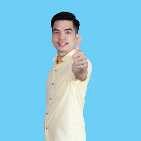 Portrait handsome young asian man wearing a yellow shirt showing thumbs up sign isolated on blue background. Asian man people. business success concept.の写真素材