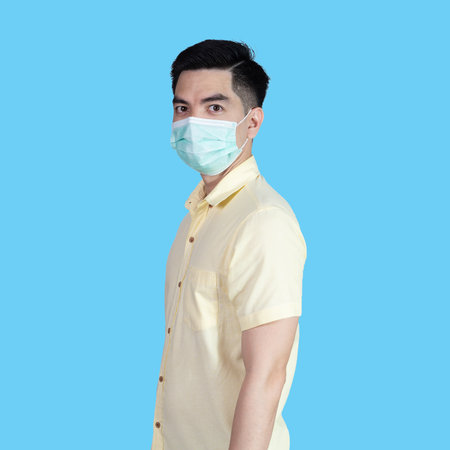 COVID-19 Coronavirus portrait handsome young asian man wearing yellow shirt and mask protection from covid 19 isolated on blue background in studio. Asian man people. COVID-19 concept.の写真素材