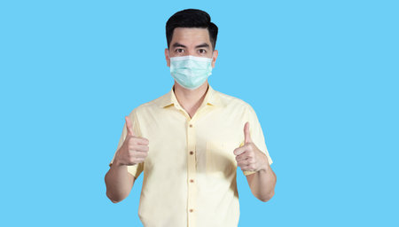 COVID-19 Coronavirus portrait handsome young asian man wearing yellow shirt and mask protection from covid 19 and thumbs up sign isolated on blue background in studio. Asian people. COVID-19 concept.の写真素材