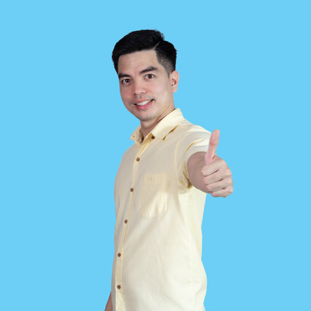 Portrait handsome young asian man wearing a yellow shirt showing thumbs up sign isolated on blue background. Asian man people. business success concept.の写真素材