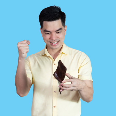 Portrait Handsome young asian man wearing a yellow shirt feeling happy money in the wallet isolated on blue background. Businessman concept. Asia people.の写真素材