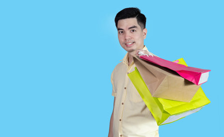 Portrait handsome young asian man wearing yellow cheerful glad content guy carrying new things colorful packages on blue background in studio. Asian man people.の写真素材