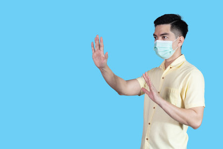 STOP! COVID-19 Coronavirus portrait handsome young asian man wearing yellow shirt and mask protection from covid 19 isolated on blue background in studio. Asian man people. COVID-19 concept.の写真素材