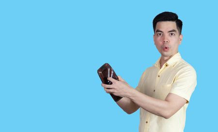 Portrait Handsome young asian man wearing a yellow shirt surpise  money in the wallet isolated on blue background. Businessman concept. Asia people.の写真素材