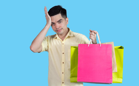 Portrait handsome young asian man wearing yellow stressful content guy carrying new things colorful packages on blue background in studio. Asian man people.の写真素材