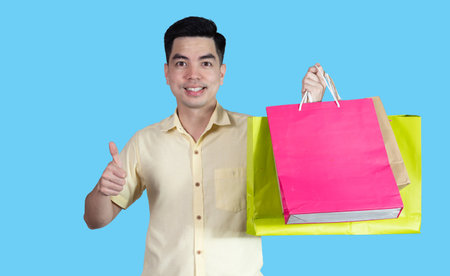 Portrait handsome young asian man wearing yellow cheerful glad content guy carrying new things colorful packages on blue background in studio. Asian man people.の写真素材