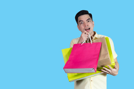 Portrait handsome young asian man wearing yellow cheerful glad content guy carrying new things colorful packages on blue background in studio. Asian man people.の写真素材