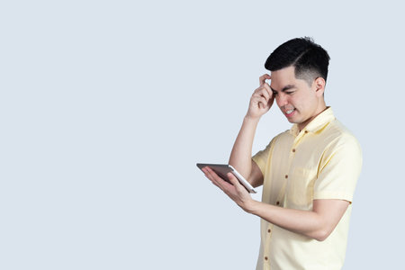 Terrible headache. portrait handsome young asian man wearing a yellow shirt holding smart phone or tablet feeling stressful isolated on white background. Businessman concept. Asia people.の写真素材