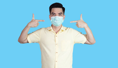 COVID-19 Coronavirus close up handsome young asian man wearing yellow shirt holding tablet and mask protection from covid 19 isolated on white background in studio. Asian man people.の写真素材