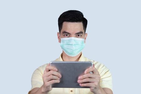 COVID-19 Coronavirus close up handsome young asian man wearing yellow shirt holding tablet and mask protection from covid 19 isolated on white background in studio. Asian man people.の写真素材