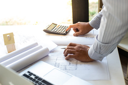 The engineer designed the house structure in the blueprint and working in the architect's room, There are equipment for designing laptops, pens, pencils, drawings And sample house with moldの写真素材