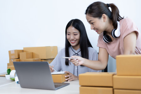 Two young Asian women chat with bright smiles to customers who shop online via laptop and confirm...