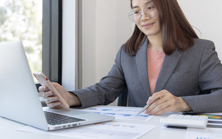 Asian female employees are analyzing market data via smartphones and have laptops to calculate the company's finances In a private office at home, Working at home and new normal concept.の写真素材
