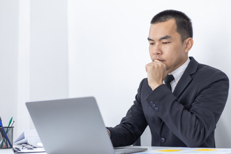 Young Asian office worker presses he finger on a laptop keyboard to analyze financial data and summarize her work in a private office, Office workers, handsome male employee in a black suit.の写真素材