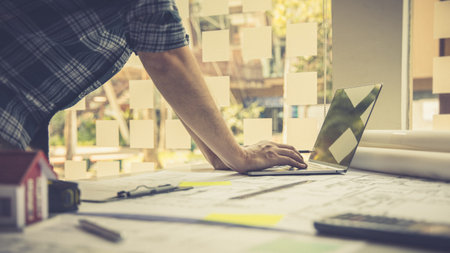 Architectural building design and construction plans with blueprints, Young man was designing a building or architecture with a ruler, pen, pencil, tape measure, architect hat and other equipment on the desk.の写真素材