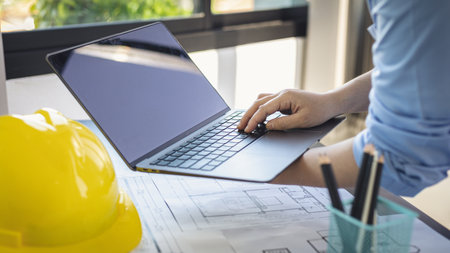 Young man with a laptop plotting a system of building structures in blueprints, Architects or engineers are designing buildings using computers to calculate the physical structure to be correct.の写真素材