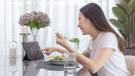 Asian woman eating fresh salad and using tablet at her home dining table, Vegetable salads are rich in vitamins and minerals, Fat-low-calorie and high-fiber diets, Healthy food, Appetizer.の写真素材