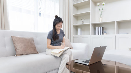 Asian woman taking notes in notebook while studying online in laptop at home, video chat, Online...